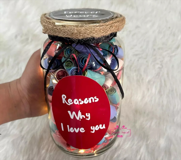 A glass jar filled with colorful rolled paper notes, featuring a red oval label that reads "Reasons Why I Love You," decorated with twine and a black ribbon.
